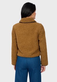 Hnědá strukturovaná fleecová bunda s límcem a kontrastním tmavým lemováním, spárovaná s modrými džínovými kalhotami, pohled zezadu.