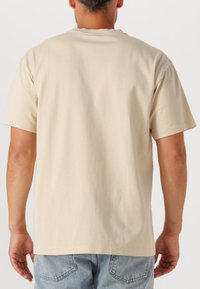 Beżowa bawełniana koszulka z krótkim rękawem, okrągłym dekoltem i prostym wykończeniem na dole, widziana z tyłu. Noszona z jasnoniebieskimi dżinsami.