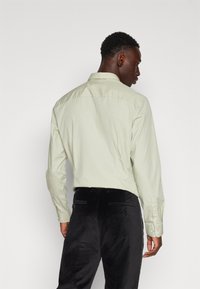 Chemise vert clair à manches longues avec un col, coutures visibles et poignets à boutons, associée à un pantalon noir ajusté avec poches arrière.
