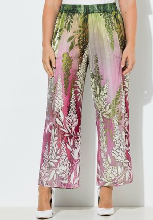 Donna che indossa pantaloni a gamba larga con stampa floreale verde, rosa e bianca abbinati a scarpe con tacchi alti bianche, davanti a una parete bianca.