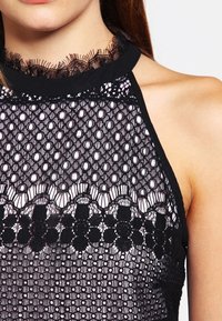 Top in pizzo nero con motivi intricati, caratterizzato da un collo alto e bordi scallop. Il materiale è trasparente con una finitura testurizzata.