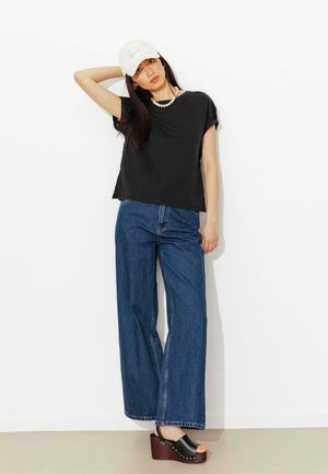 Femme portant un t-shirt noir, un jean large bleu, une casquette blanche, un collier de perles et des sandales à plateforme noires, debout contre un mur blanc.