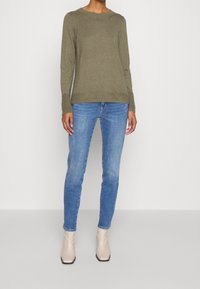 Olivengrüner Pullover mit Rundhalsausschnitt, langen Ärmeln, kombiniert mit schmal geschnittenen blauen Jeans und beigen Stiefeletten. Glatte Textur und lässiges Design.