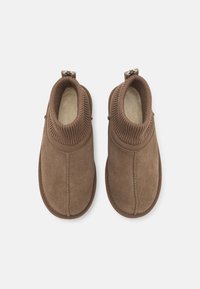 Chaussons en daim marron avec un revers en tricot, orteil arrondi et détails cousus. Comprend une languette pour un enfilage facile. Doublure intérieure douce.