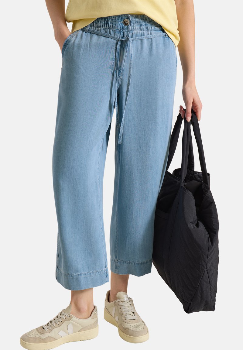 Personne portant un pantalon large bleu clair court avec une taille élastique et tenant un sac cabas matelassé noir.