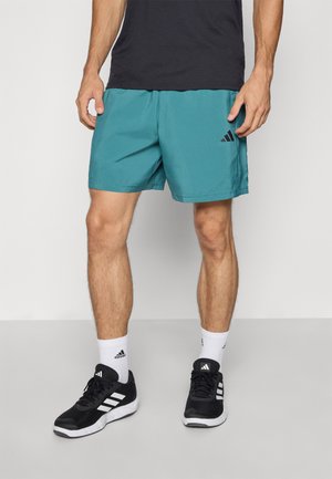 Uomo che indossa pantaloni sportivi teal, scarpe nere Adidas, calze bianche Adidas e una maglietta nera, in piedi contro uno sfondo chiaro e semplice.