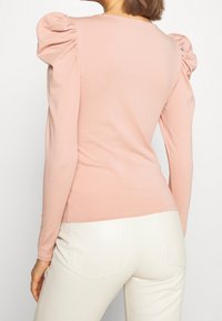 Långärmad topp i ljusrosa med puffärmar, mjukt material och figurnära silhuett, kombinerad med vita tajta byxor.