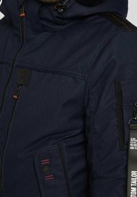 Giacca imbottita blu navy con tessuto testurizzato, vari tasche, dettagli neri e cuciture rosse a contrasto sulla tasca sinistra.