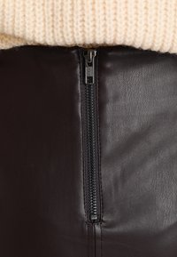 Brauner Lederrock mit einem zentralen Reißverschlussverschluss und einem metallischen Ziehgriff, ergänzt durch einen strukturierten Strickpullover darüber.