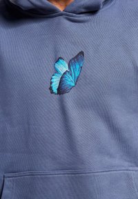 Sudadera azul con un gráfico de mariposa azul y negra en el pecho. La tela tiene un acabado texturizado y un bolsillo tipo canguro.