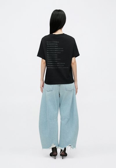 T-shirt preta de algodão com design de texto nas costas, combinada com jeans azul claro oversize com bainhas desfiadas e bolsos laterais. Sapatos de salto pretos.