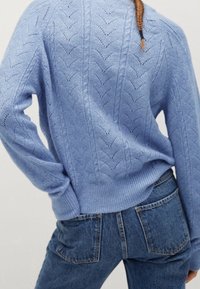Žena oblečená v svetle modrém pleteném svetru s vzorovanými otvory a modrých džínech, stojící s jednou rukou na boku.