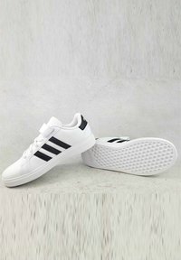 Sneakers atletici bianchi con strisce nere, colletto imbottito, materiale sintetico liscio e suola in gomma bianca testurizzata. Cinturino regolabile con velcro.
