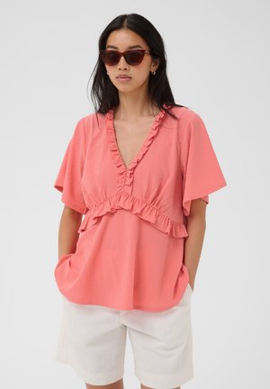 Femme portant un chemisier ample corail rose à volants et col en V, short blanc et lunettes de soleil foncées, debout les mains dans les poches devant un fond uni.