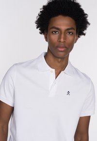 Camiseta polo blanca con una tapeta de dos botones, textura de algodón suave y logo bordado en azul marino en el lado izquierdo del pecho. Manga corta y corte clásico.