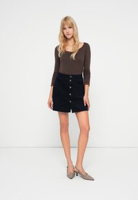 Giovane donna con capelli biondi lunghi che indossa una maglia marrone a maniche lunghe, una gonna nera con bottoni frontali e tacchi beige a punta, in piedi contro un muro bianco.