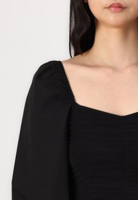 Haut noir présentant un décolleté carré, des manches bouffantes froncées et un tissu légèrement texturé. Le design met en valeur un corsage ajusté.