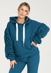 Turkusowa bluza z kapturem wykonana z miękkiego materiału. Posiada marszczone rękawy, kieszeń z przodu oraz kontrastowe białe sznurki do regulacji.