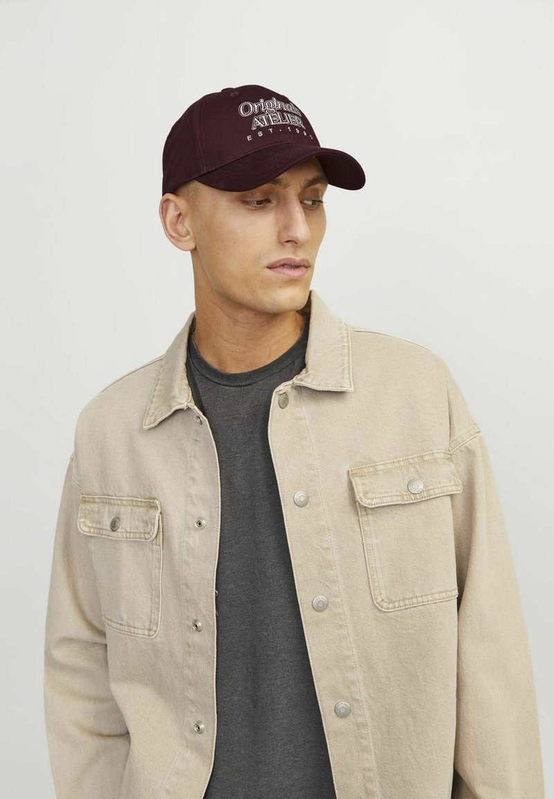 Gorra de béisbol color burdeos con el logotipo bordado "Original Atelier", combinada con una chaqueta de mezclilla beige claro y una camiseta gris oscuro debajo.