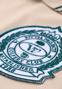 Beiger Stoff mit einem kreisförmigen, bestickten Aufnäher, der ein grünes Logo, Lorbeerblätter, den Text "Riviera Project Golfers Club" und "Gegründet 1988" zeigt.