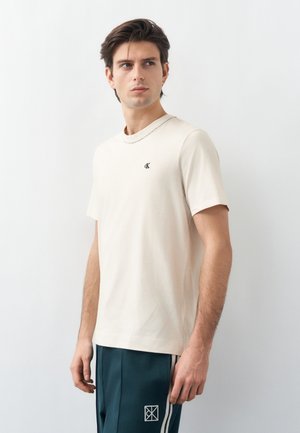 Νέος άνδρας με σκούρα μαλλιά, ντυμένος με μπεζ μπλούζα Calvin Klein και σκούρο πράσινο παντελόνι με λευκές ρίγες, στέκεται μπροστά σε έναν λευκό τοίχο.