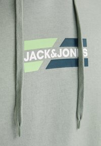 Haut à capuche vert clair avec cordons de serrage, arborant un logo graphique en blanc, bleu foncé et accents vert clair. Texture de tissu douce.