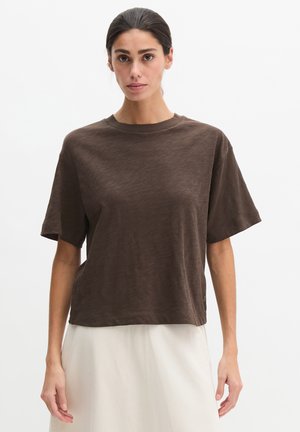 Femme aux cheveux foncés attachés, portant une chemise marron à manches courtes et ample, ainsi qu'une jupe de couleur claire, debout devant un fond uni.