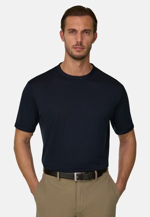 Mężczyzna nosi zwykły czarny T-shirt z krótkim rękawem włożony w beżowe spodnie z czarnym paskiem, stoi z rękami w kieszeniach.