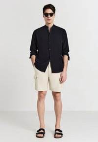 Chemise noire à manches longues avec fermeture à boutons, shorts cargo beige jusqu'aux genoux et sandales noires, debout contre un fond uni.