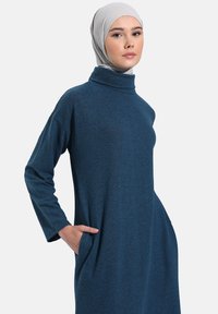 Abito lungo color teal con maniche lunghe e colletto alto finto, vestibilità comoda, tasche laterali e tessuto testurizzato. La modella indossa un foulard grigio chiaro.