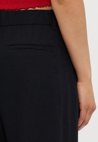 Zbliżenie na talię i biodra osoby w czarnych spodniach z tylną kieszenią oraz w czerwonym topie na krótkim rękawie, ręka spoczywa obok ciała.