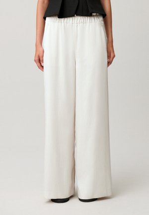 Donna che indossa pantaloni bianchi a vita alta, a gamba larga con vita elastica, abbinati a un top nero e scarpe nere.