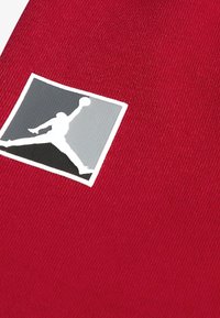Tessuto rosso con una finitura texturizzata presenta un logo Jumpman bianco e grigio in un bordo quadrato, posizionato nell'angolo in alto a sinistra.