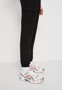 Bílé sportovní boty se stříbrnými a růžovými akcenty, síťovanou svrchní částí a gumovou podrážkou. Spárováno s černými tepláky se bočními kapsami.