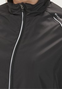 Chaqueta negra con cremallera y cuello alto, con una textura suave y detalle en blanco a lo largo de la cremallera. Logotipo de la marca situado en el hombro.
