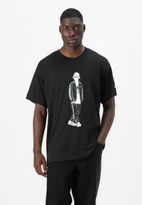 Μαύρη μπλούζα με γραφικό ενός ανάλαφρα ντυμένου ατόμου με καπέλο. Χαλαρή εφαρμογή, κοντά μανίκια, φτιαγμένη από μαλακό βαμβακερό υλικό.