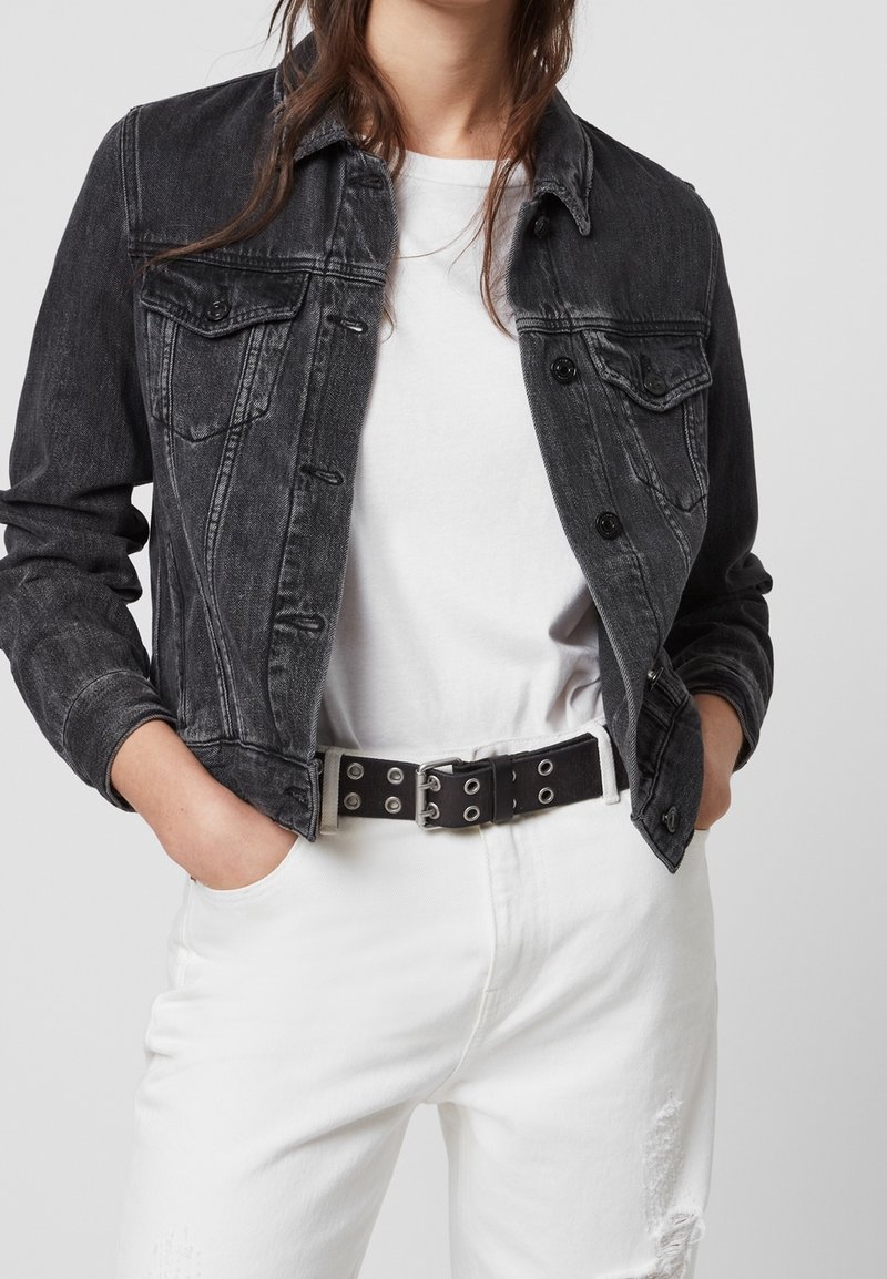 Černá džínová bunda s klasickým střihem, zapínáním na knoflíky vepředu a kapsami na hrudi. Bílé tričko pod ní, doplněné o bílé zničené džíny a černý opasek.