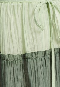 Vestido de tela texturizada en verde claro, con un dobladillo fruncido en verde oscuro, cintura ceñida y cinturón de lazo. Diseño de bloques de color horizontal.