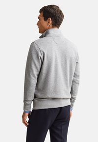 Homme de dos portant un pull gris zippé à col montant et un pantalon sombre sur un fond uni.