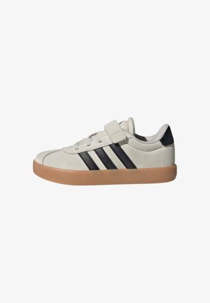 Baskets basses beige avec des rayures noires, une semelle en caoutchouc couleur beige foncé, des lacets et une bande velcro sur fond blanc.