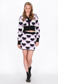 Completo in maglia a due pezzi composto da un cardigan corto e una gonna aderente. Base lavanda con motivi a cuore neri e dettagli neri. Stivali neri.