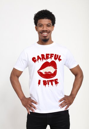 Hvid bomulds t-shirt med et iøjnefaldende grafik af røde læber med hugtænder og dryppende tekst, der siger "PAS PÅ JEG BIDDER" i rødt.