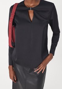 Haut noir avec découpe en forme de goutte, manches longues et texture lisse. Associé à une jupe en cuir noir ajustée et une bretelle d'épaule rouge.