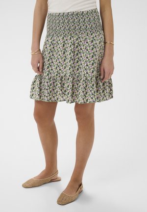 Femme portant une jupe verte à imprimé floral avec une taille smockée et un ourlet à volants, associée à des chaussures slingback tissées beige et des bracelets dorés.