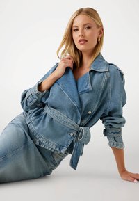 Giacca in denim di colore azzurro chiaro, design oversize con ampie rever, vita con cintura e dettagli a bottoni. Tessuto testurizzato e maniche arrotolate.