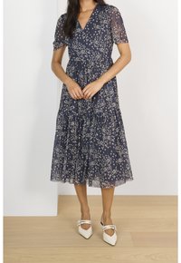 Navyblaues Blumenkleid mit kurzen Puffärmeln, gestuftem Design und cremefarbenem Blumenmuster. Kombiniert mit spitzen weißen Schuhen.