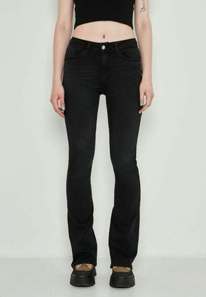 Osoba ubrana w czarne dzwony, krótki czarny top i buty na platformie stojąca na tle jasnego, jednolitego tła.