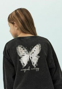 Sweat-shirt en charbon avec une texture douce, arborant un grand graphique de papillon blanc et la phrase "énergie magique" dans une police décorative.