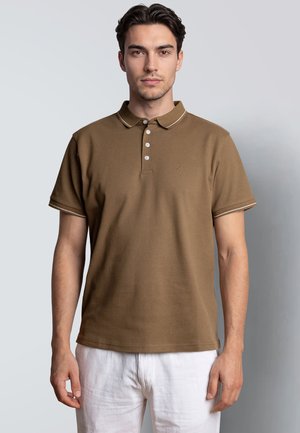 Jonge man met donker haar, gekleed in een bruin poloshirt met korte mouwen en witte broek, kijkt recht vooruit tegen een effen lichte achtergrond.