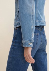 Persona vistiendo una chaqueta de denim azul claro y unos pantalones de tiro alto azul oscuro, con la mano relajada a su lado contra un fondo neutro.